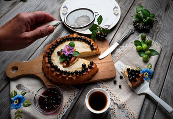 Big round cheese cake with black currant and a cup of coffee on a wooden grey table.