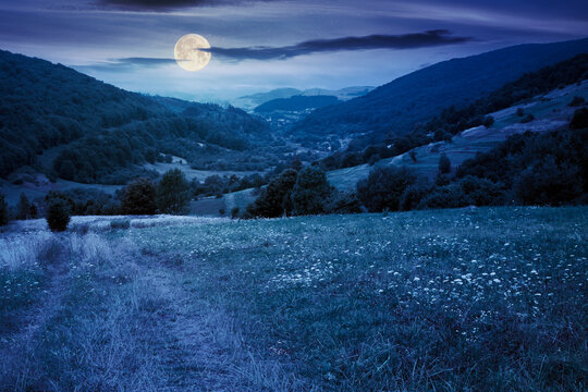 country road through rural field at night. suburban summer landscape in mountains in full moon light. village in the distant valley. cloudy day
