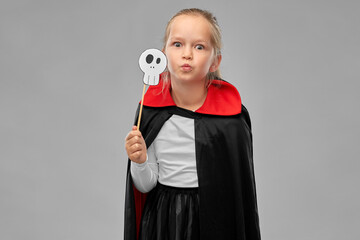halloween, holiday and childhood concept - girl in dracula costume with black cape holding scull...