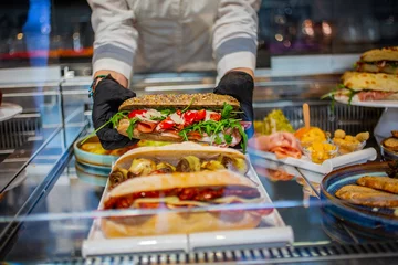 Foto op Plexiglas Banconista mentre sistema un panino integrale con pomodori, bresaola, rucola e scaglie di parmigiano in vetrina © Vincenzo VAD