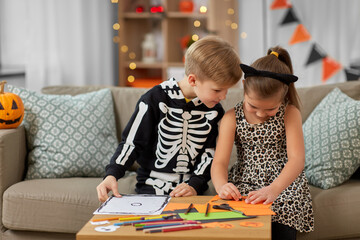 Fototapeta premium halloween, holiday and childhood concept - smiling little boy and girl in costumes doing crafts at home