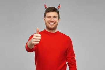 Fotobehang holiday, theme party and people concept - happy smiling man in halloween costume of devil showing thumbs up over grey background © Syda Productions