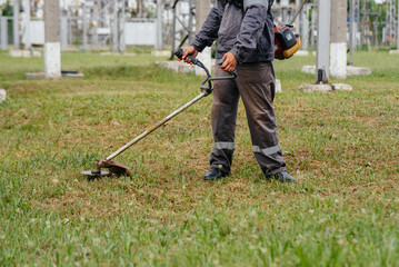 A young man mowing grass on the territory of an electric substation in overalls. Grass cleaning at the enterprise, implementation of fire safety measures