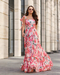 Elegant woman in long pink summer dress with floral print walking at city street. Outdoor full...