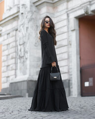 Fototapeta premium Young beautiful woman with brunette hair wearing long black dress and sunglasses, holding leather handbag and walking at city street on a summer day. Outdoor full length fashion portrait. Female model