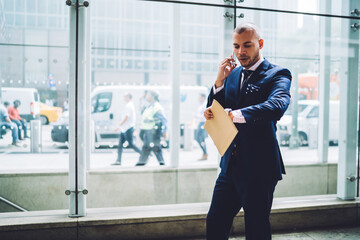 Serious businessman in elegant suit checking time hurrying in office talking on phone with colleague,handsome busy executive manager walking with documents arranging meeting during mobile conversation