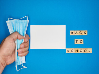 On blue background, close-up of hand clutching medical mask and phrase made of wooden letters, distance learning, copy space. the concept of returning to school after coronavirus pandemic.