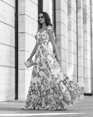 Elegant woman in long pink summer dress with floral print walking at city street. Outdoor full length portrait. Pretty female model