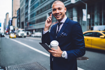 Portrait of positive executive manager in elegant suit standing on road calling in taxi service,...
