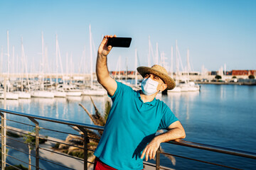 Tourist with a mask and shade taking a selfie in a marina with leisure boats in the background.