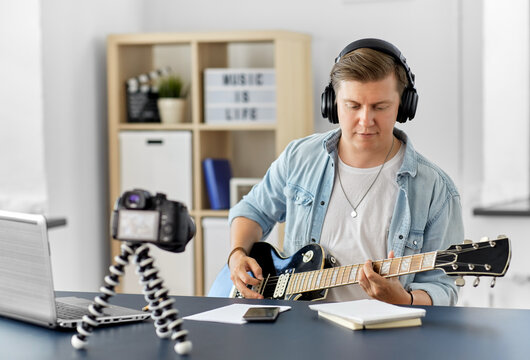 video blogging, music and people concept - young man or musician in headphones with camera videoblogging and playing guitar sitting at table at home