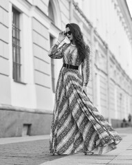 Pretty female model in gray dress with stripes and sunglasses standing and posing at city street on a summer day. Full length fashion oudoor portrait