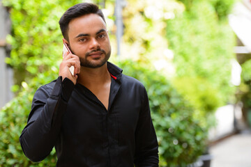 Young handsome bearded Indian businessman talking on the phone at the park