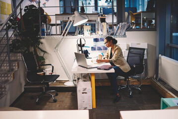 Young female talented administrative manager working with financial documents and reports organizing them into folder in database using computer and wireless connection in company headquarter indoor