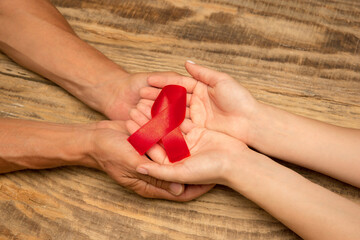 Female and male hands holding red HIV and AIDS awareness ribbon isolated on wooden background. Concept of healthcare and medicine, worldwide supporting, volunteer campaign against illness.