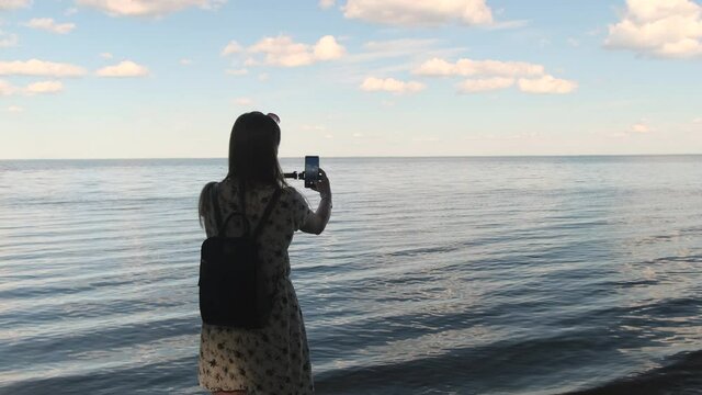 Back view portrait of woman making selfie in the background of beautiful seascape using a smartphone and the monopod at sunset, slow motion
