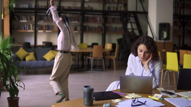 Open public library. Happy dreadlocks beautiful girl in white blouse and beige pants funny dancing on background while her friend concentrated on her studying, sitting at the table works with laptop