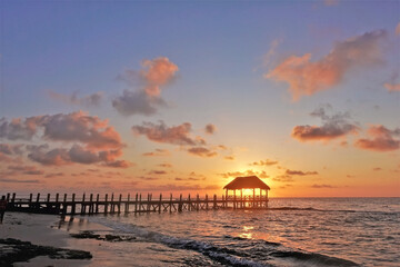 Sunrise over the Caribbean Sea. Waves roll on the beach sand. Above the water there is a wooden...