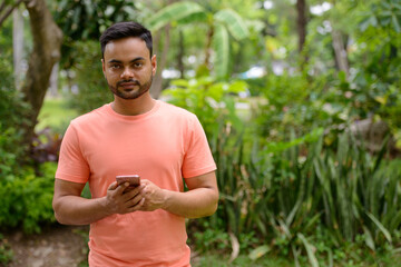 Naklejka na ściany i meble Young handsome bearded Indian man using phone at the park
