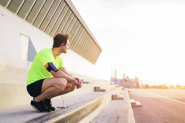 Young sports man thoughtful looking away while having a rest after workout training outdoors in urban setting, male runner listen to music in headphones though smart phone and admiring morning sunrise