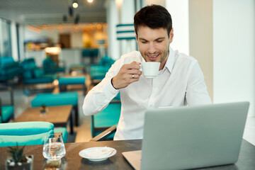 Smiling businessman drinking coffee while using computer in a cafe.