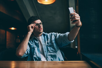 Handsome hipster guy in spectacles for vision correction making selfie photos for attracting followers in social networks, young man posing and taking pictures via front camera on mobile phone
