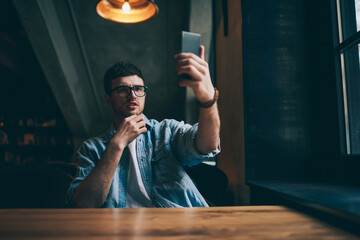 Handsome hipster guy in eyeglasses making video conversation with colleague calling by application on modern smartphone device, young man looking in web camera on cellular phone and clicking selfie