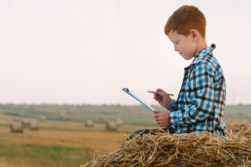 Naklejka na ściany i meble Little farmer on top of a straw bale holds a tablet with a letter and a pen in his hands on a background of hay bales
