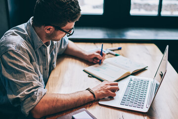 Casual dressed hipster guy writing organisation plan in textbook for education sitting at desktop with netbook,young man writing essay in notebook and preparing for academy test with modern technology - obrazy, fototapety, plakaty