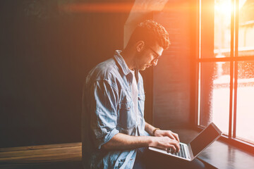 Caucasian male freelancer sitting near cafeteria window and typing new article in modern laptop application, young man blogger browsing wireless internet and updating profile in social networks