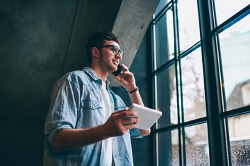 Successful hipster guy making positive conversation with friend while downloading file on digital tablet, smiling man in trendy spectacles calling via app on mobile phone using 4g internet indoors