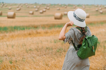 Beautiful girl in a hat and with a backpack in the middle of a wheat field with bales in a summer evening