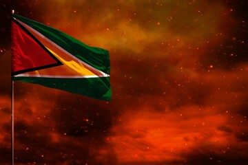 Fluttering Guyana flag mockup with blank space for your text on crimson red sky with smoke pillars background. Troubles concept.