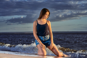 Fototapeta na wymiar A young brunette girl in a black T-shirt and denim shorts kneels on the sandy shore near the water's edge. Summer sunny evening.
