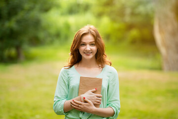 Smiling redhead girl posing with book in hands, enjoying reading outdoors