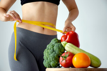Slim woman measuring her waist size with tape measure and set of fresh vegetables on cutting board in defocus. Balanced nutrition, dieting or detox concept.