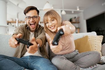 Husband and wife playing video game with joysticks in living room. Loving couple are playing video games at home.. - obrazy, fototapety, plakaty