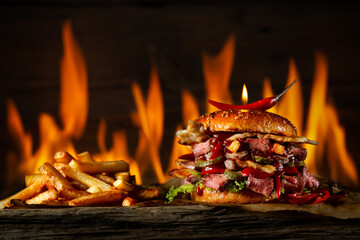 Spicy burger with beef and red chili and French fries lies on the table against the background of fire