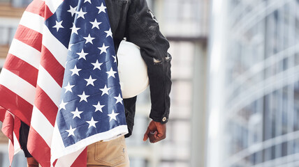 View from behind. Patriot holding USA flag. Conception of pride and freedom. Young african american man in black jacket outdoors in the city standing against modern business building - Powered by Adobe
