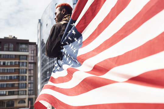 View from behind. Patriot holding USA flag. Conception of pride and freedom. Young african american man in black jacket outdoors in the city standing against modern business building