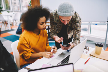 Bearded designer showing design application installed on smartphone to africa american colleague sitting at laptop computer in office for productive work of developing creative project