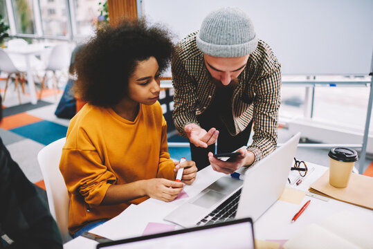 Bearded designer showing design application installed on smartphone to africa american colleague sitting at laptop computer in office for productive work of developing creative project
