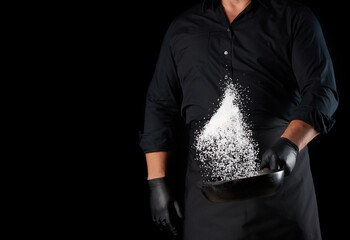man in a black uniform holding a round cast iron pan with salt, the chef tosses white salt up