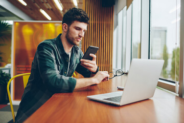 Fototapeta premium Confident young man freelancer checking email on smartphone while working at modern laptop computer using wireless internet connection.Male blogger updating app on cellular sitting at netbook device