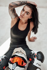 Fototapeta na wymiar Beautiful looking brunette girl with tatoos in a black and red tracksuit, white boots and with a motorcycle helmet, posing in a white photo studio, sitting on the floor and holding her hair with her