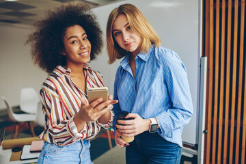 Portrait of cheerful diverse friends in casual wear smiling at camera while updating profile in social networks and messaging in online chat on modern smartphone using high speed internet connection