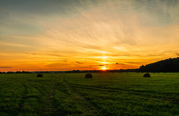 Scenic view at beautiful sunset in green shiny field with hay stacks, bright cloudy sky, country road and golden sun rays with glow, summer valley landscape