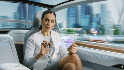 Attractive Senior Female Reading News on a Futuristic Transparent Tablet Computer with Augmented Reality Interface while Sitting on a Backseat of Autonomous Car. Self-Driving Van Rides on Public Road.