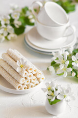 Delicate sweet cookies in the form of tubes in coconut flakes filled with vanilla cream on a white plate and a yellow pear with two tea mugs in the background. 