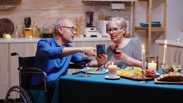 Man in whellchair using smartphone sitting at the table in the kitchen while romantic dinner. Imobilized paralyzed handicapped elderly husband scolling on phone enjoying the festive male.